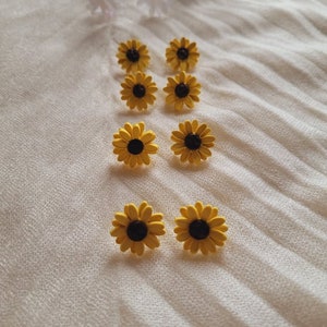 May include: Eight yellow sunflower earrings with black centers. The earrings are arranged in two rows of four on a white fabric background.