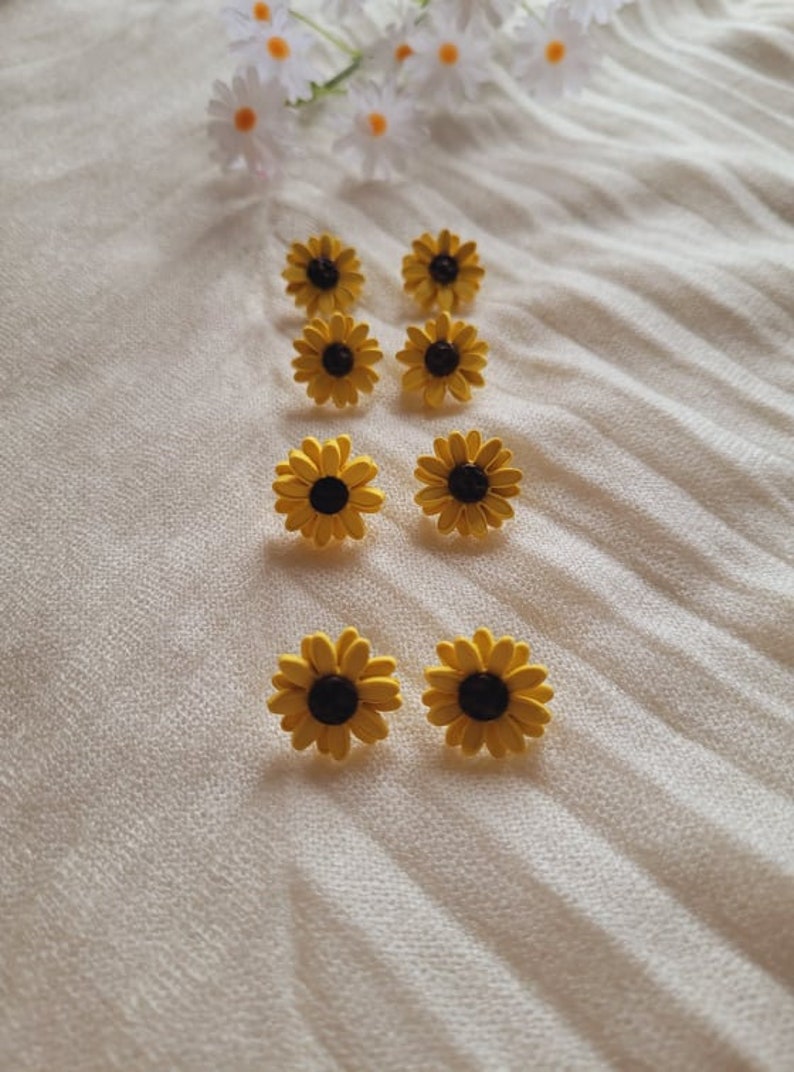 May include: Eight yellow sunflower earrings with black centers. The earrings are arranged in two rows of four on a white fabric background.