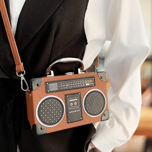 Puede incluir: Bolso bandolera marrón con forma de radiocasete vintage. El bolso presenta un asa plateada, detalles de altavoces y un diseño de dial de radio. La palabra "BOOH" está impresa en la esquina superior derecha del bolso.