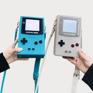 May include: Two handheld game console-shaped crossbody bags. One is light blue with a black screen and buttons, the other is off-white with a gray screen and burgundy buttons. Both have the text "GAME BAG BAIDA".