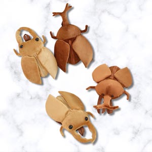 May include: Four plush insect toys in shades of brown and tan are arranged on a white marble background. The toys include two beetles with large, open wings, one with a zipper, and two other insect designs with various horn and leg details.