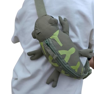 May include: A novelty crossbody bag shaped like a cartoon frog. The bag is olive green with lime green accents and a zipper closure. The frog has outstretched arms and legs, and a long strap.