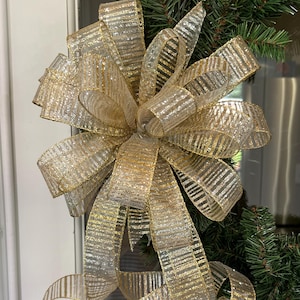 May include: A large, decorative bow made of sheer, gold-striped ribbon. The bow is attached to a wreath of green pine needles. The ribbon has a metallic sheen, and the bow is prominently displayed, suggesting festive decor.