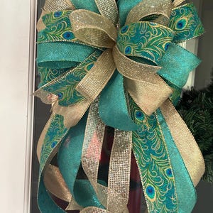 May include: A large decorative bow featuring teal and gold ribbons. The teal ribbons are adorned with a peacock feather pattern. Gold ribbons are layered throughout the bow, creating a festive and elegant design. The bow is likely for a wreath or other holiday decoration.