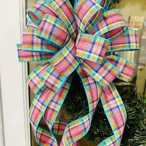May include: A large, colorful plaid bow with a turquoise trim. The bow is made of a vibrant fabric in shades of pink, orange, yellow, green, and blue. It is attached to a green wreath.