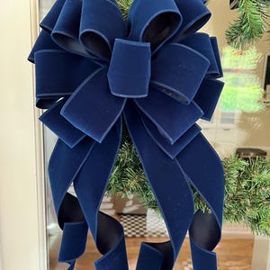 May include: A large, multi-looped decorative bow crafted from deep navy blue velvet ribbon. The bow features a central knot with multiple symmetrical loops radiating outward and two long, flowing ribbon tails that curl at the ends.
