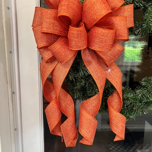 May include: A large, multi-looped decorative bow made from orange, textured fabric ribbon. The bow features a central knot with several voluminous loops and two long, curling tails that hang downward, attached to a dark green pine wreath.