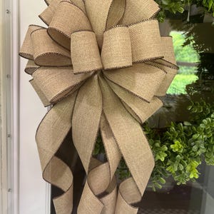 May include: A large, decorative bow made of tan burlap ribbon with dark brown stitched edges. The bow is attached to a wreath with green foliage. The bow is hanging on a white door.