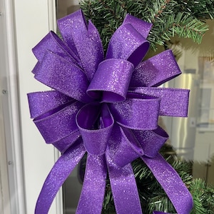May include: A large, decorative purple bow with a glittery finish. The bow is attached to a green garland, creating a festive look. The bow's ribbons are looped and curled, adding dimension and visual interest to the holiday decor.