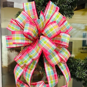 May include: A large, decorative bow made of colorful plaid ribbon. The ribbon features a pattern of pink, green, yellow, and blue squares. The edges of the ribbon are trimmed in pink. The bow is attached to a green wreath.