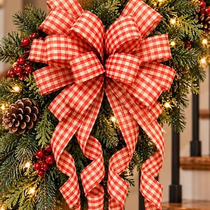 May include: A large decorative bow made of red and white gingham ribbon attached to a lush evergreen garland. The garland is adorned with clusters of small red berries, brown pinecones, and warm white string lights on a wooden post.