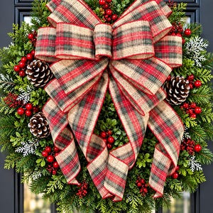 May include: A circular holiday wreath featuring dense green evergreen foliage, natural brown pinecones, and clusters of bright red berries. A large, prominent bow made of red, green, and tan plaid fabric with wired edges is centered on the wreath.