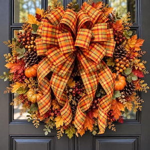 May include: A decorative autumn wreath featuring a large central bow made of orange, yellow, and green plaid ribbon. The wreath is adorned with artificial maple leaves, pinecones, small orange pumpkins, and clusters of red and yellow berries.