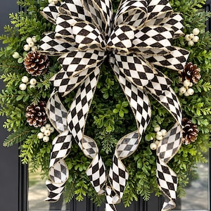 May include: A round wreath made of green boxwood foliage, decorated with brown pinecones and clusters of small white berries. A large, multi-looped bow made of black and white diamond-patterned ribbon with gold edges is centered on the wreath.