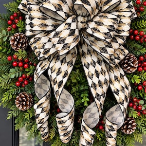 May include: A decorative wreath featuring lush green evergreen foliage, clusters of bright red berries, and brown pinecones. A large, multi-looped bow made of cream-colored ribbon with a black and gold diamond harlequin pattern is centered.