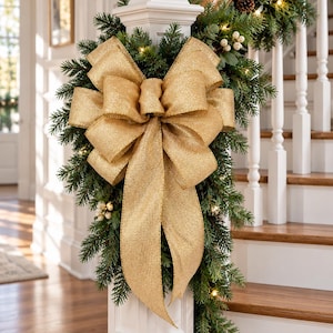 May include: A large, shimmering gold fabric bow with a textured finish attached to a lush green pine garland. The garland is adorned with small white berries, pinecones, and warm white string lights, draped over a white wooden staircase banister.