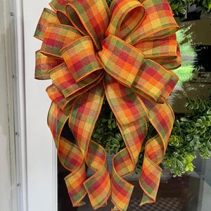 May include: A large decorative bow with a plaid pattern in fall colors of orange, yellow, and green. The bow is attached to a green wreath, creating a festive autumn decoration.