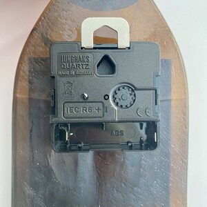 Puede incluir: Un mecanismo de reloj Junghans Quartz negro con un colgador de metal plateado est&aacute; fijado a una botella de vidrio marr&oacute;n. El mecanismo del reloj tiene el texto "MADE IN GERMANY" y "IEC R6 +" impreso.
