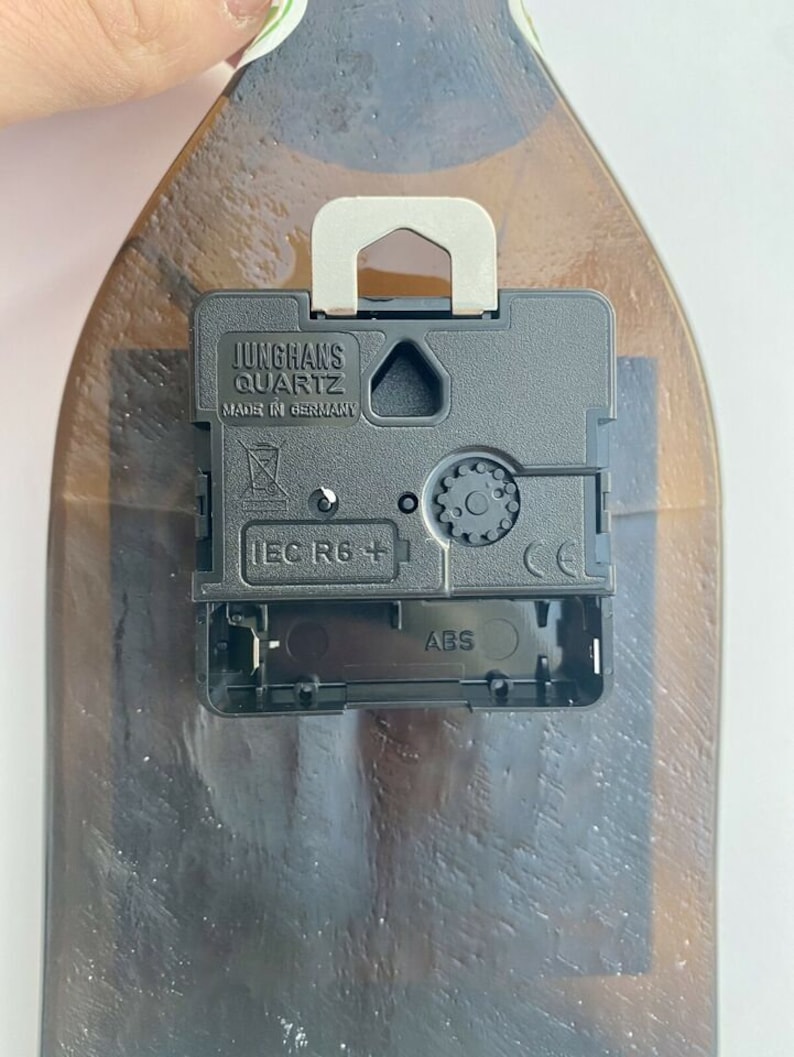 Puede incluir: Un mecanismo de reloj Junghans Quartz negro con un colgador de metal plateado est&aacute; fijado a una botella de vidrio marr&oacute;n. El mecanismo del reloj tiene el texto "MADE IN GERMANY" y "IEC R6 +" impreso.