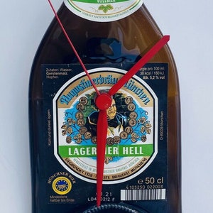 Puede incluir: Una botella de cerveza marr&oacute;n con una etiqueta verde y blanca que dice "HELLES VOLLBIER". La etiqueta tambi&eacute;n incluye el texto "Lager Hell" y "Augustiner-Br&auml;u M&uuml;nchen". Una esfera de reloj est&aacute; superpuesta.