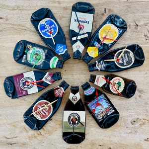 May include: A collection of clocks made from recycled beer bottles. Each clock features a different beer label and has gold or red clock hands. The bottles are arranged in a circular pattern on a wooden surface.