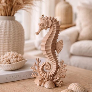 May include: A beige seahorse sculpture with coral and seashells on a wooden table. The seahorse is intricately detailed and stands upright. The words "Coastal Living" are visible on a book in the background. The overall theme is coastal decor.