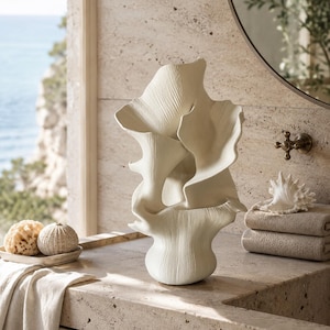 May include: A sculptural off-white ceramic vase with a textured, organic, and flowing form that resembles layered coral or sea flora. The surface features fine vertical ridges and a matte finish, standing upright with multiple undulating petal-like sections.