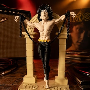 Peut inclure: Une figurine d'un homme torse nu aux cheveux noirs, portant un pantalon noir et une ceinture dorée, debout devant une arche beige. La figurine a des chaînes noires attachées aux poignets et est fixée sur un socle avec le texte The Doors en relief.