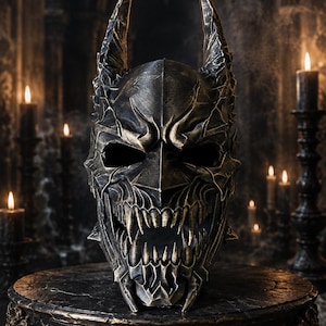Könnte beinhalten: Ein dunkler Helm mit metallischer, verwitterter Oberfläche, der spitze, längliche fledermausartige Ohren und eine bedrohliche, skelettartige Gesichtsmaske mit freiliegenden, gezackten Zähnen aufweist. Die Oberfläche hat eine texturierte Bronze-Schwarz-Patina.