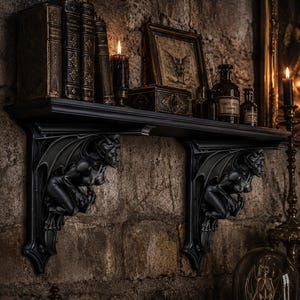 Peut inclure: Une étagère murale noire soutenue par deux supports sculptés identiques représentant des gargouilles ailées. L'étagère contient plusieurs livres anciens, une bougie noire allumée, une illustration de chauve-souris et des flacons d'apothicaire.