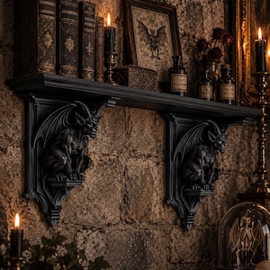 Peut inclure: Une étagère murale noire soutenue par deux supports en forme de gargouilles détaillées. L'étagère contient plusieurs livres anciens avec des lettres dorées, une illustration de chauve-souris encadrée, des flacons en verre et une bougie noire allumée.