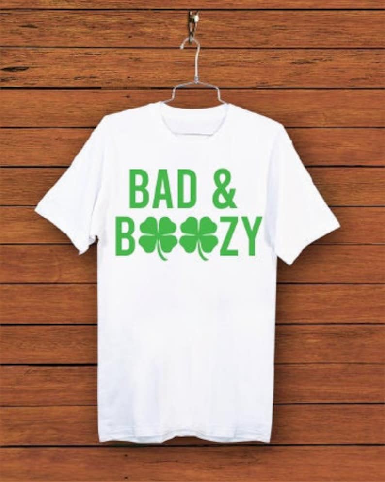 K&ouml;nnte beinhalten: Wei&szlig;es T-Shirt mit den Worten "BAD & BOOZY" in gr&uuml;nen Blockbuchstaben und gr&uuml;nen Kleeblatt-Grafiken. Das Shirt h&auml;ngt an einem Metallb&uuml;gel vor einem Holzgrund.