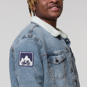 May include: A light blue denim jacket with a white sherpa collar and a patch on the left sleeve. The patch features a mountain range and a bicycle in a navy blue color.