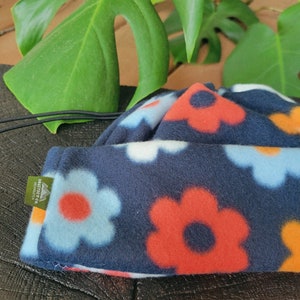 May include: A navy blue fleece hat with a floral pattern in red, light blue, white, and orange. The hat has a drawstring and a small tag. The hat is resting on a dark, textured surface.