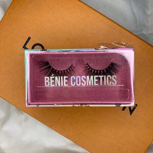 Könnte beinhalten: Nahaufnahme einer pinkfarbenen und weißen Schachtel mit zwei Paaren künstlicher Wimpern. Die Schachtel hat eine glitzernde Oberfläche und den Text "BÉNIE COSMETICS" aufgedruckt.