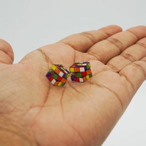 Puede incluir: Dos pendientes pequeños de forma cuadrada con un patrón de cuadrícula multicolor que recuerda a un cubo de rompecabezas clásico. Cada pendiente muestra una vibrante disposición de cuadrados rojos, azules, amarillos, verdes, naranjas y blancos.
