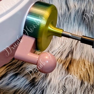 May include: A close-up of a pink and white knitting loom with a green and gold yarn winder attached. The winder has a metal rod that extends out from the side. The loom and winder are sitting on a fluffy, brown and white faux fur surface.