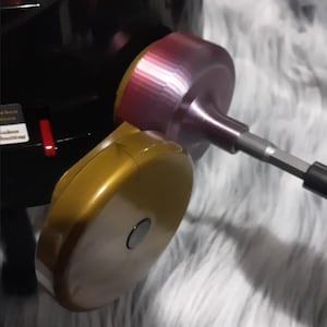 May include: Close-up of a black and gold drill with a pink and silver drill bit attached. The drill is on a white fuzzy surface.