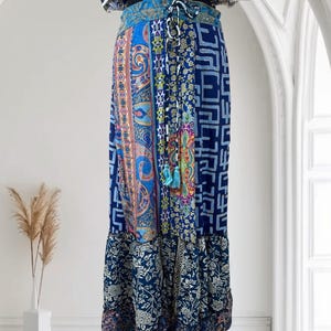 May include: A long, flowing skirt with a patchwork design in shades of blue, featuring paisley, geometric, and floral patterns. The skirt has a drawstring waist and a ruffled hem, creating a bohemian aesthetic.