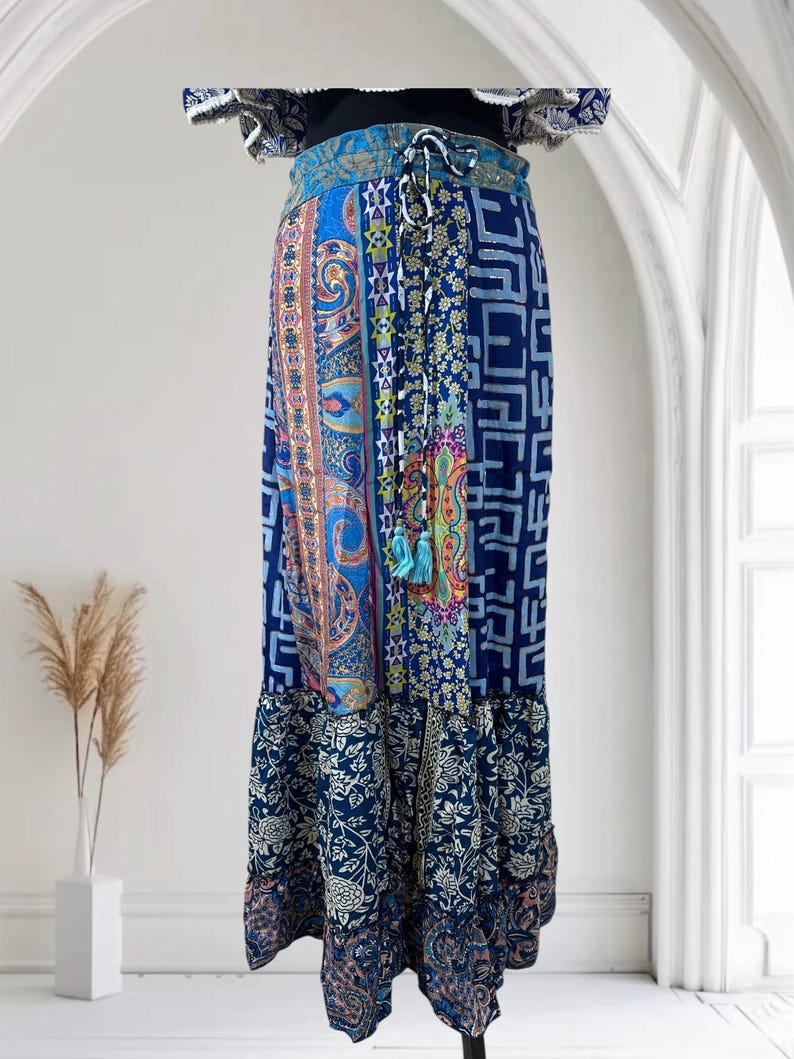 May include: A long, flowing skirt with a patchwork design in shades of blue, featuring paisley, geometric, and floral patterns. The skirt has a drawstring waist and a ruffled hem, creating a bohemian aesthetic.