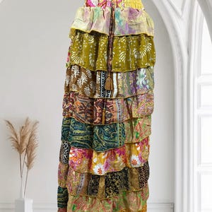Op de afbeelding: Een lange, gelaagde rok met een verscheidenheid aan kleurrijke stoffen met patronen. De rok heeft een tailleband met trekkoord en een kwastdetail. De lagen bevatten bloemen-, paisley- en abstracte ontwerpen in tinten groen, geel, roze en bruin.