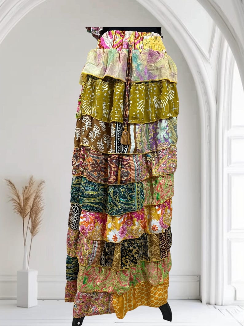 Op de afbeelding: Een lange, gelaagde rok met een verscheidenheid aan kleurrijke stoffen met patronen. De rok heeft een tailleband met trekkoord en een kwastdetail. De lagen bevatten bloemen-, paisley- en abstracte ontwerpen in tinten groen, geel, roze en bruin.
