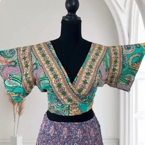 May include: A colorful wrap top and skirt set. The top features a V-neck and short sleeves with a teal, pink, and yellow paisley pattern. The skirt has a gathered waist and a mix of purple, blue, and orange floral prints. The outfit is displayed on a black mannequin.
