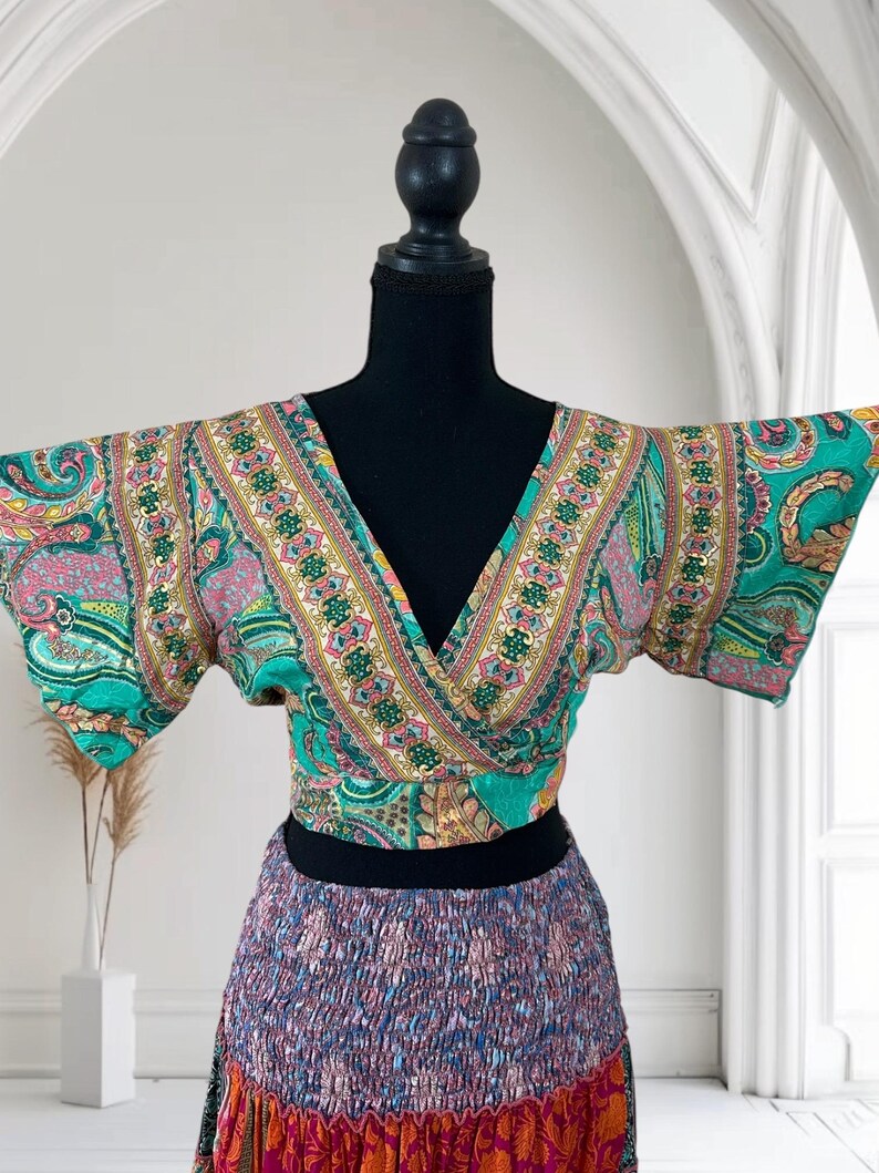 May include: A colorful wrap top and skirt set. The top features a V-neck and short sleeves with a teal, pink, and yellow paisley pattern. The skirt has a gathered waist and a mix of purple, blue, and orange floral prints. The outfit is displayed on a black mannequin.
