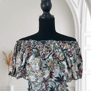 May include: Off-the-shoulder top with a floral pattern in shades of gray, red, and yellow. The top has short sleeves and an elasticated waist. The fabric is gathered at the neckline and waist, creating a ruffled effect. The top is displayed on a black mannequin.