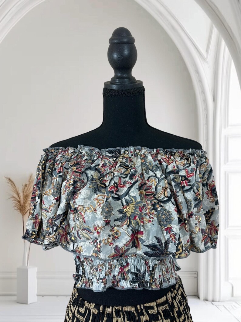 May include: Off-the-shoulder top with a floral pattern in shades of gray, red, and yellow. The top has short sleeves and an elasticated waist. The fabric is gathered at the neckline and waist, creating a ruffled effect. The top is displayed on a black mannequin.