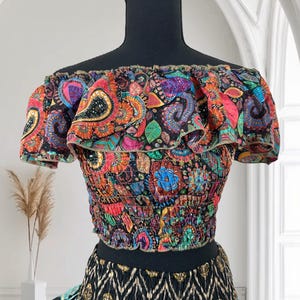 May include: A colorful off-the-shoulder crop top with a paisley pattern in shades of red, orange, blue, and green. The top has a ruffled neckline and short sleeves. The bottom of the top is elasticized. The skirt below is black with a gold and teal pattern.