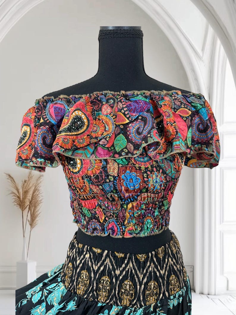 May include: A colorful off-the-shoulder crop top with a paisley pattern in shades of red, orange, blue, and green. The top has a ruffled neckline and short sleeves. The bottom of the top is elasticized. The skirt below is black with a gold and teal pattern.