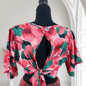 May include: A floral print top with a keyhole back and tie closure. The top features a pink and red floral pattern with green leaves. The sleeves are short and flared. The top is displayed on a black mannequin.