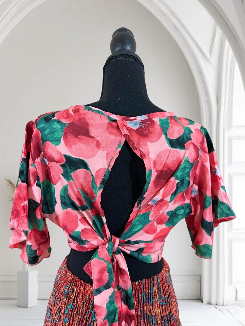 May include: A floral print top with a keyhole back and tie closure. The top features a pink and red floral pattern with green leaves. The sleeves are short and flared. The top is displayed on a black mannequin.